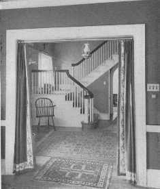 This attractive Colonial hallway shows a good arrangement
of rugs. The border on the porti&egrave;res spoils the effect, but the lamp is
well chosen.