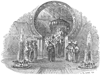 A man walks, head bowed, through an arch, with three men flanked on either side of it,
while other men are in a group behind him